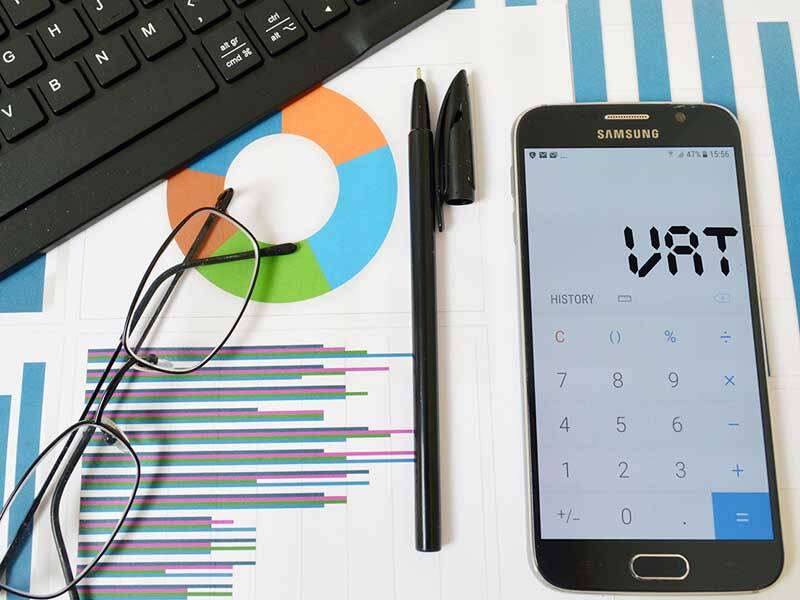 Picture of charts and information under spectacles, a pen, and a mobile phone with calculator app open that shows VAT on the screen