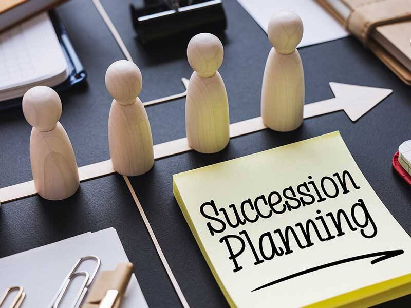 Wooden figures lined up in line with a paper arrow, with Succession Planning written on a postitpad, along with other stationary and objects
